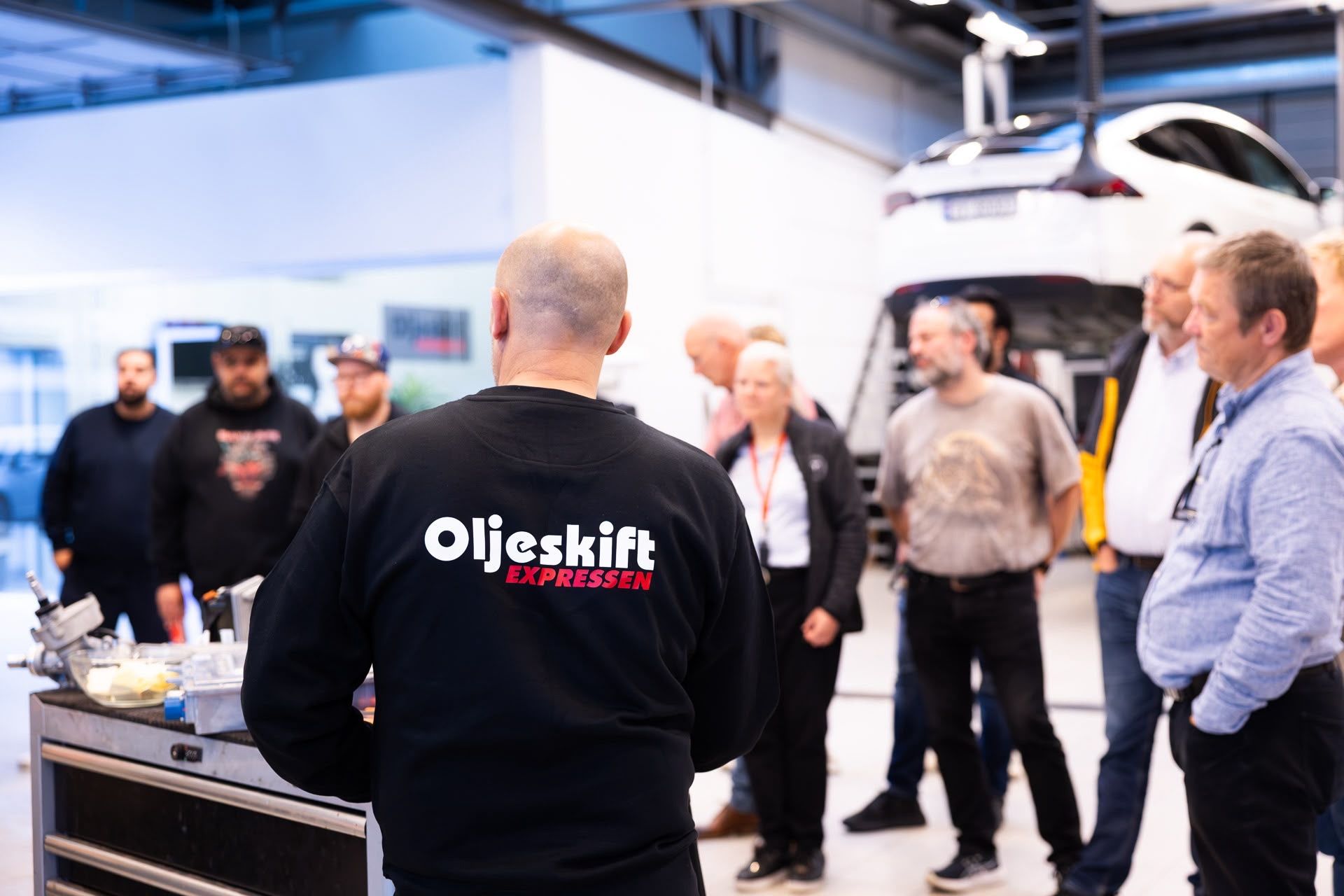 Technician in an Oljeskiftexpressen sweater showing parts to visitors, Tesla on the lift behind