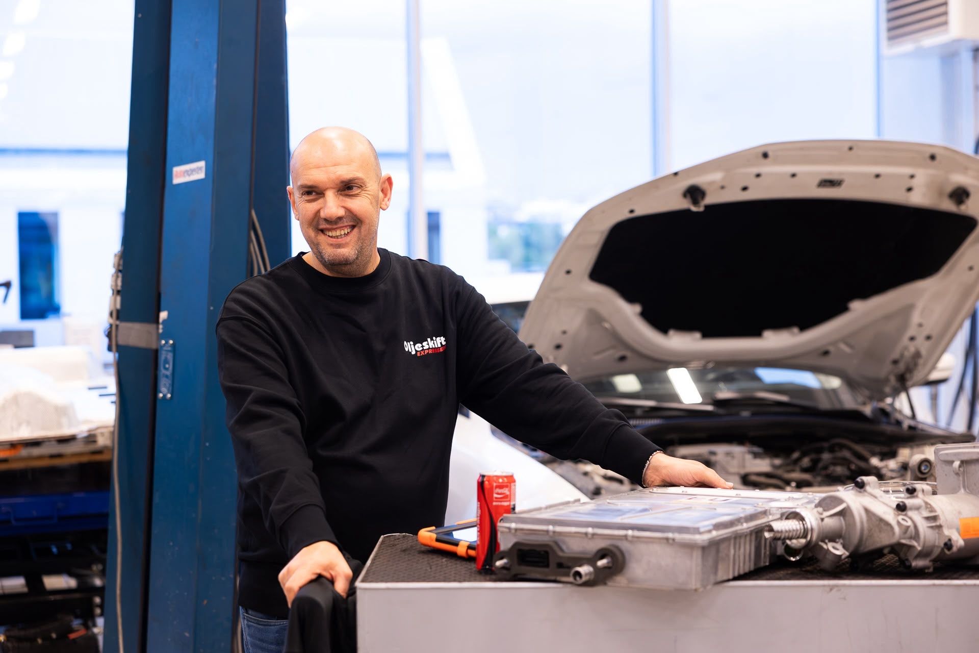 Smiling technician at a car with the hood open and a drive unit on the workbench